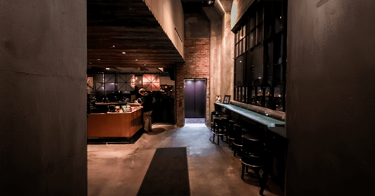 Interior of a stylish, moody retrofit space, likely a restaurant or bar, showing a compact LU/LA elevator entrance with a dark door blended into a brick wall, demonstrating how the lift integrates into older buildings.