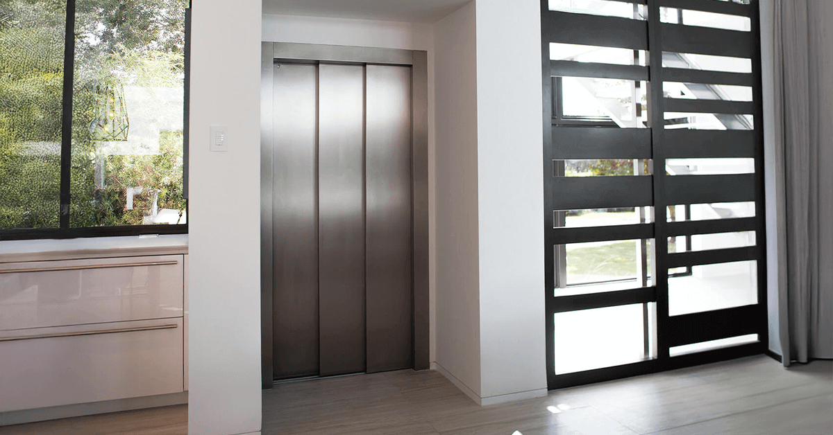 Modern, sleek stainless steel home elevator doors built seamlessly into a white wall of a residential interior with large windows looking out onto greenery, highlighting a compact and elegant household elevator design.