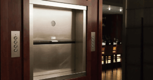 A stainless steel dumbwaiter car and landing doors set within dark wood paneling in a commercial setting, featuring call buttons on the jamb, showcasing a modern, professional-grade service lift.