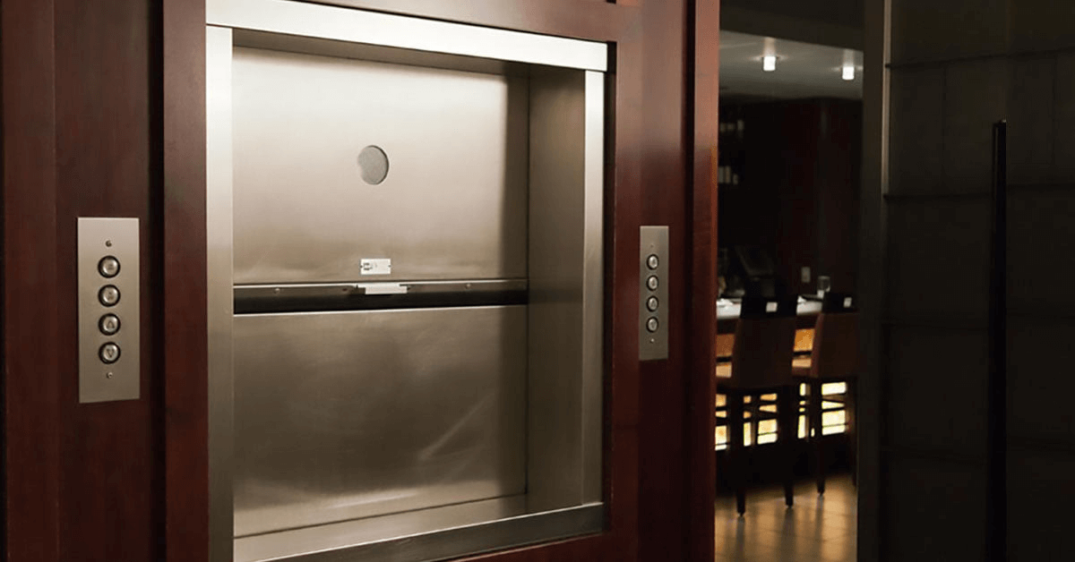 A stainless steel dumbwaiter car and landing doors set within dark wood paneling in a commercial setting, featuring call buttons on the jamb, showcasing a modern, professional-grade service lift.