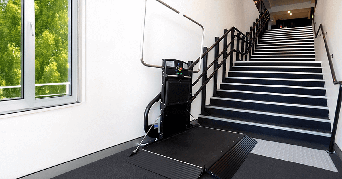 Modern black Inclined Platform Lift (IPL) or Staircase Chair Lift folded at the bottom of a dark staircase in a modern commercial building, providing an accessibility solution that is an alternative to a home elevator or domestic lifts.