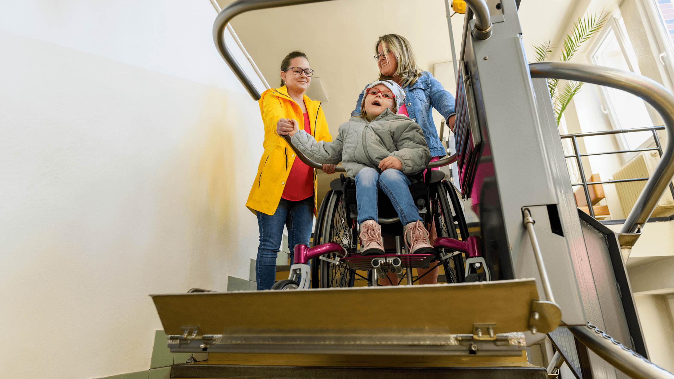 Two women assisting a young girl in a wheelchair on an inclined platform lift moving up a staircase.
