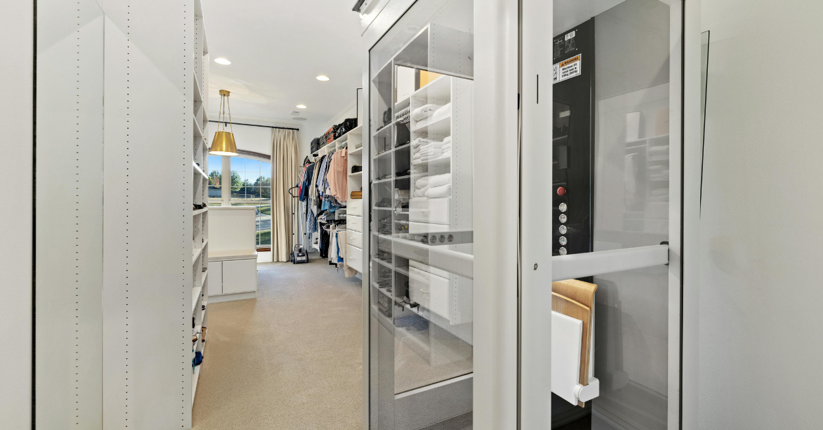 Modern home elevator with a clear glass door integrated seamlessly into a spacious white luxury walk-in closet.