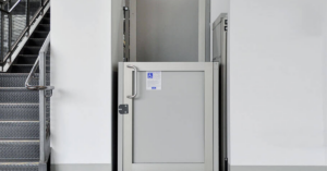 Silver commercial vertical platform lift with an accessibility sign installed next to a set of metal indoor stairs.