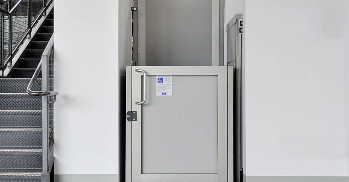 Silver commercial vertical platform lift with an accessibility sign installed next to a set of metal indoor stairs.